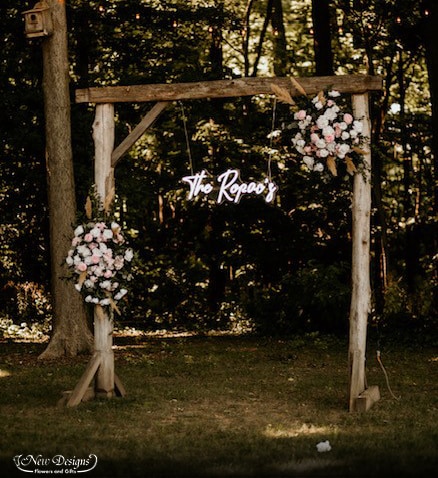 A Square Arch in front of a forest, With Installments of White and Blush flowers, and a Light up Neon Sign hanging from the Top Bar